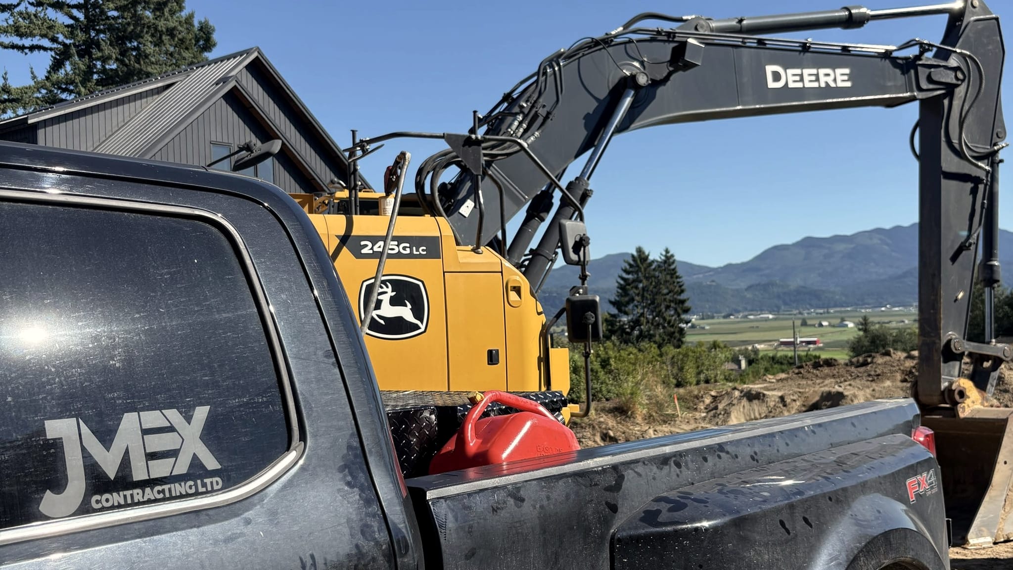 A JMEX Contracting truck and a John Deere excavator on a Fraser Valley job site, mountains behind