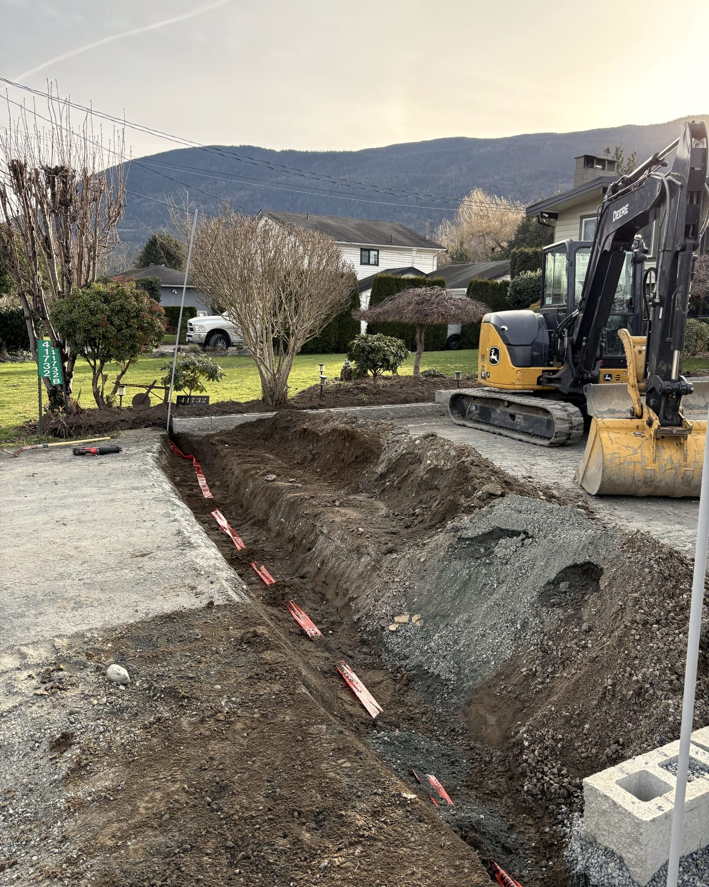 A John Deere excavator and a freshly dug trench on a Fraser Valley residential property, mountains behind